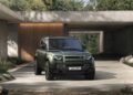 A green Land Rover Defender Refreshed: Here's What’s New For 2026, parked in front of a modern concrete house surrounded by greenery and rocks.