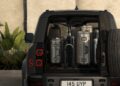 A black Land Rover Defender Refreshed for 2026 sits with its rear door open, revealing a bicycle and three silver suitcases neatly packed in the cargo area.