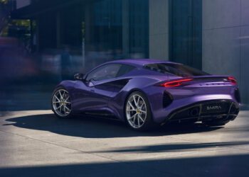 A purple Lotus Emira sports car is parked on a city street beside a modern building, with blurred figures in the background.