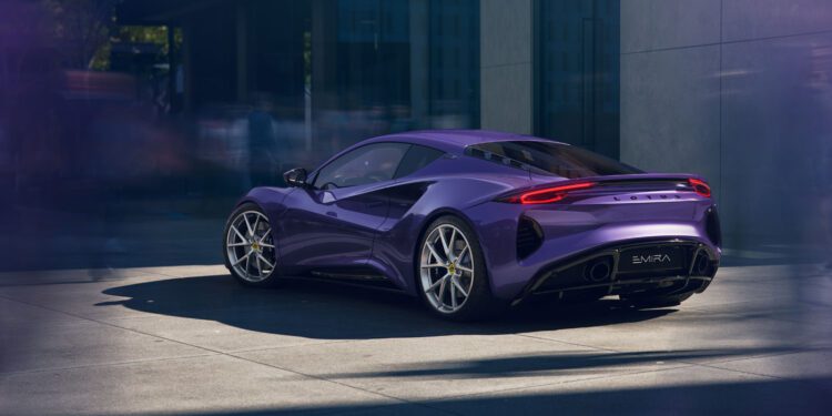 A purple Lotus Emira sports car is parked on a city street beside a modern building, with blurred figures in the background.