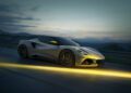 A silver Lotus Emira sports car driving at dusk on a winding road, illuminated by a yellow light streak on the pavement.