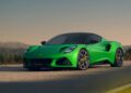 A bright green Lotus Emira sports car is parked on a paved road with a scenic, blurred landscape in the background under a clear sky.