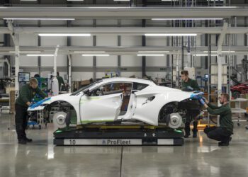 Three workers assemble a white Lotus sports car on a factory floor, with the car body elevated and wheels not yet attached—a glimpse into U.S. production at the crossroads of innovation and precision craftsmanship.