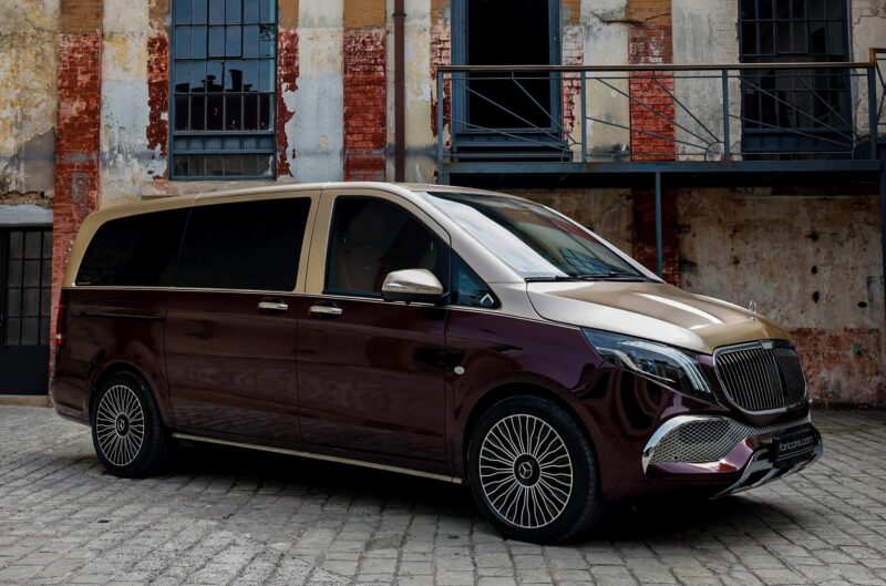 A luxury minivan with a two-tone maroon and gold exterior—a unique creation—is parked on cobblestone in front of a weathered brick building.