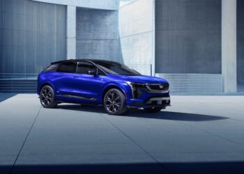 A blue Cadillac Lyriq electric SUV is parked on a concrete surface in front of a modern building with grey walls.