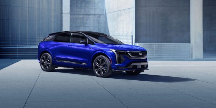 A blue Cadillac Lyriq electric SUV is parked on a concrete surface in front of a modern building with grey walls.