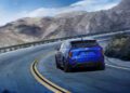 A blue 2026 Optiq-V SUV with 519HP drives along a winding mountain road with guardrails, surrounded by rocky hills under a blue sky.
