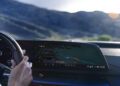 A person drives a Cadillac 2026 Optiq-V, glancing at a wide digital dashboard screen displaying navigation with a map and route guidance.