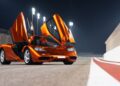 An orange sports car with its butterfly doors open is parked on a racetrack at night under bright lights, reminiscent of the ultra-exclusive 1-of-1 McLarens featured in the historic sale of 20 rare models.