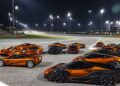 A group of orange McLaren sports cars, part of the 20 Ultra-Exclusive 1-of-1 McLarens being offered in a historic sale, parked on a racetrack under bright stadium lights at night.