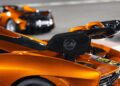 Close-up of the carbon fiber rear wing on an orange sports car with "LM" emblem; another is blurred in the background on a racetrack—part of the 20 ultra-exclusive 1-of-1 McLarens being offered in a historic sale.