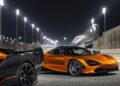 Two orange McLaren sports cars are parked on a racetrack at night under bright stadium lights, part of the 20 ultra-exclusive 1-of-1 McLarens being offered in a historic sale, with a grandstand and fencing visible in the background.