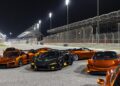 A group of orange and black McLaren sports cars, part of the 20 ultra-exclusive 1-of-1 McLarens being offered in a historic sale, are parked on a race track at night, with grandstands and track lights in the background.