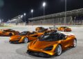Several orange McLaren sports cars, part of the 20 ultra-exclusive 1-of-1 McLarens being offered in a historic sale, are parked on a racetrack at night under bright stadium lights with grandstands in the background.
