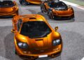 Three orange McLaren supercars are parked on a racetrack surface, including the legendary F1 in front, as part of the 20 ultra-exclusive 1-of-1 McLarens being offered in a historic sale.