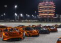A group of orange and black McLaren sports cars, part of the 20 ultra-exclusive 1-of-1 McLarens being offered in a historic sale, are parked on a racetrack at night with a brightly lit round building and stadium lights in the background.