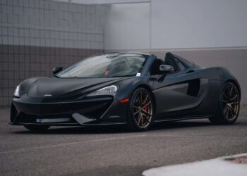 A matte black convertible sports car, perfect for those who love stealth mode, is parked in an outdoor lot near a building with tiled and concrete walls.