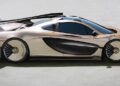 A gold and black McLaren P1 with a low profile, aerodynamic design, large rear wing, and unique wheel rims—echoing Frank Stephenson’s visionary touch—is parked on a light-colored surface.