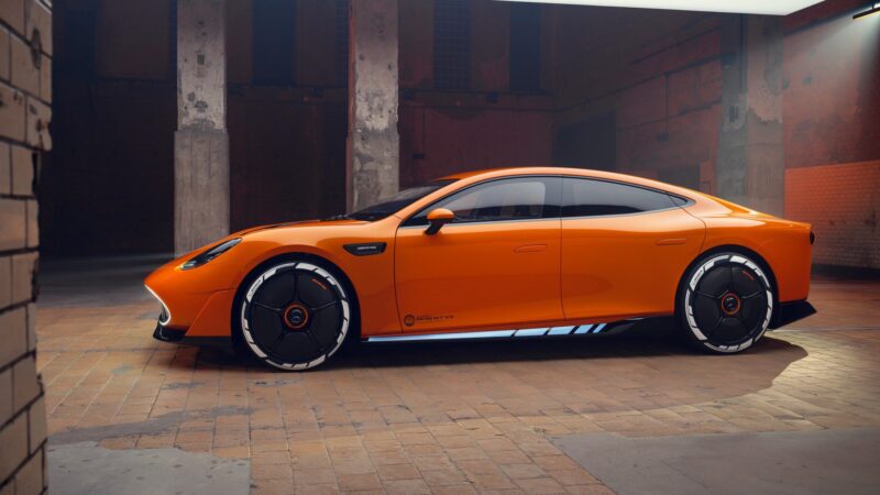 A sleek, orange, four-door Mercedes sports car with black and white accents—possibly inspired by the AMG GT XX Concept 341-HP EV—is parked indoors on a tiled floor under industrial lighting.