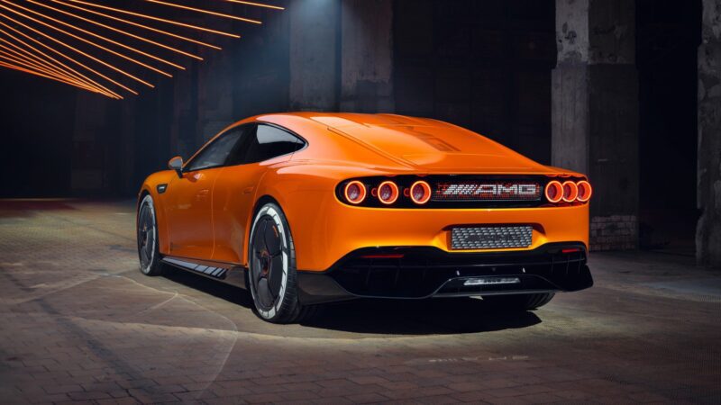 An orange Mercedes AMG GT XX Concept luxury sports car with AMG badging is parked in an industrial space with exposed beams and illuminated ceiling lights.