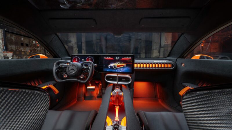 Interior of a modern Mercedes AMG GT XX sports car featuring two displays, a unique steering wheel, orange accent lighting, carbon fiber details, and the power of a 341-HP EV.