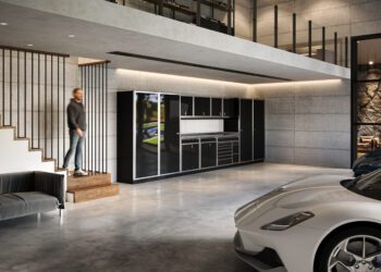 A man descends stairs into a modern garage with concrete walls, a black sofa, a kitchen area, and two parked sports cars—an impressive space that reflects refined living and luxury home accessories.
