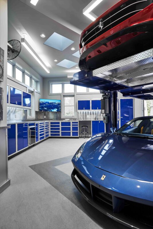 Modern garage with blue cabinets, a car lift holding a red sports car above a blue sports car, and a wall-mounted TV—perfect for those who appreciate refined living and luxury home accessories.