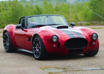 A red Cobra GT Roadster by AC Cars, featuring black racing stripes, is parked on a paved surface, with greenery and a chain-link fence in the background—a true production classic.