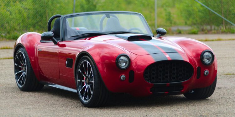 A red Cobra GT Roadster by AC Cars, featuring black racing stripes, is parked on a paved surface, with greenery and a chain-link fence in the background—a true production classic.