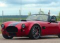 A red AC Cars Cobra GT Roadster with black racing stripes is parked on a paved surface near a chain-link fence and open landscape, ready for production thrills.