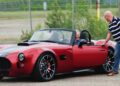Two men interact with a red Cobra GT Roadster parked in an outdoor lot near a chain-link fence; one is seated inside, while the other stands by the door, showcasing AC Cars' latest production.