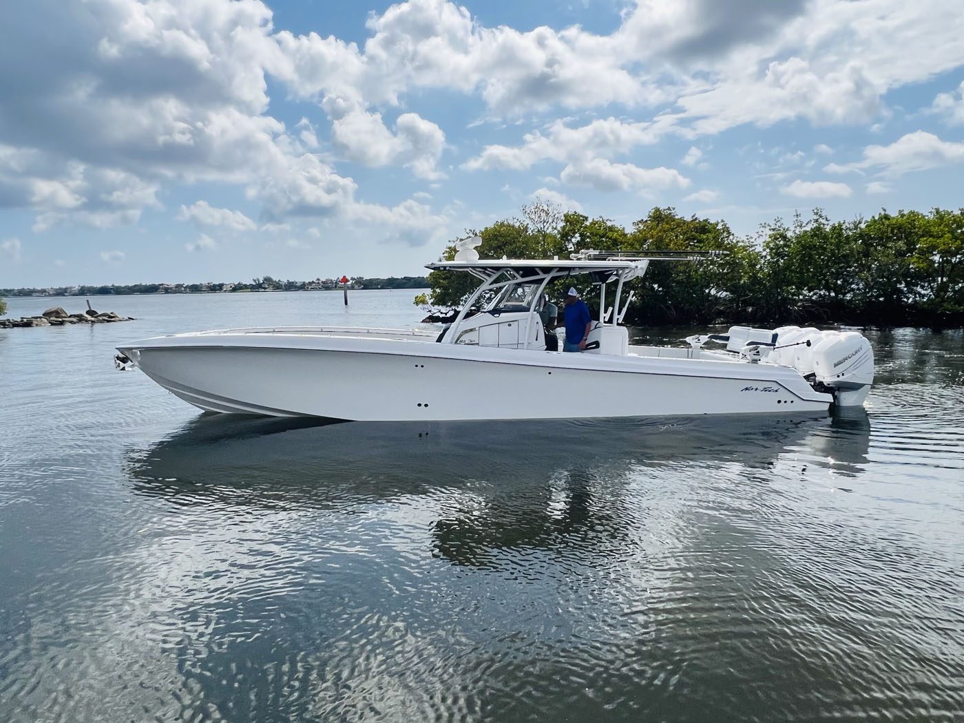 A white center console motorboat with dual outboard engines floats on calm water near a shoreline with trees under a partly cloudy sky, its hull protected by advanced Marine Protection Film for superior vessel preservation.