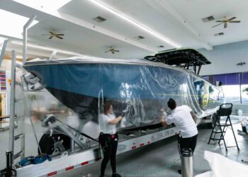 Two people apply Marine Protection Film to a large boat on a trailer inside a spacious, well-lit room with ceiling fans, ensuring optimal vessel protection and marine preservation.