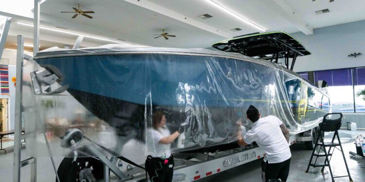 Two people apply Marine Protection Film to a large boat on a trailer inside a spacious, well-lit room with ceiling fans, ensuring optimal vessel protection and marine preservation.