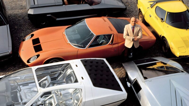 A man in a beige jacket stands among several vintage sports cars, including an orange, a yellow, and two silver vehicles—one of which is The Last Wedge: Centennial 1-of-1 Bertone Nuccio, headed to auction—parked on cobblestone.