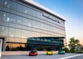 Three Porsche sports cars in red, yellow, and green are parked at One Porsche Drive in Atlanta, in front of a modern glass building with a large Porsche logo at sunset.