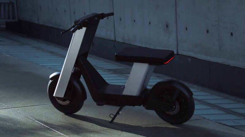A modern, black and gray Infinite Machine Olto electric scooter with a seat is parked on a sidewalk beside a concrete wall near the bike lane.