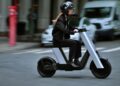 Person wearing a helmet rides a modern Infinite Machine electric scooter in the bike lane on a city street, with blurred background indicating motion.