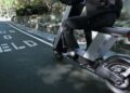 A person rides an Olto electric moped on a city bike lane marked with "PEDS TO YIELD." Trees and a stone wall line the path, capturing the freedom of the Infinite Machine experience.