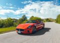 A red Maserati GT2 Stradale convertible sports car drives on a winding road surrounded by greenery under a partly cloudy sky, evoking the spirit of Motor Valley Fest.