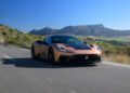 A bronze and black Maserati GT2 Stradale drives on a highway through a rural landscape with mountains in the background under a clear blue sky.