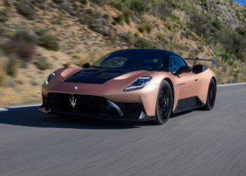 A bronze Maserati MC20 Cielo sports car drives on a paved road with hills and greenery in the background, showcasing its style at the Motor Valley Fest.
