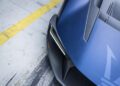 Close-up of a blue sports car’s rear corner, highlighting its taillight, carbon fiber accents, and part of a yellow line on the concrete ground.