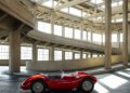 A vintage red sports car is parked inside a large, concrete, industrial-style building with ramps and geometric ceiling patterns.