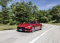A red Maserati sports car drives on a winding road with trees in the background under a blue sky, capturing the spirit of classic cars and Mille Miglia racing.