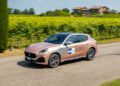 A light brown Maserati SUV with racing decals drives on a rural road beside a vineyard, evoking the spirit of historic cars from the 1000 Miglia, with a house and trees visible in the background.