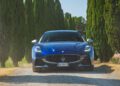 A blue Maserati sports car, reminiscent of the 1000 Miglia's historic cars, is parked on a gravel road lined with tall green cypress trees on both sides under a clear sky.