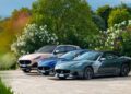 Three Maserati cars—a beige SUV, a blue coupe, and a gray convertible—are parked side by side on a paved area, surrounded by lush greenery and flowering bushes, evoking the spirit of historic cars at the 1000 Miglia.