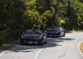 Two dark-colored sports cars, including a Maserati, drive along a winding road bordered by dense green trees on a sunny day, echoing the spirit of the 1000 Miglia.