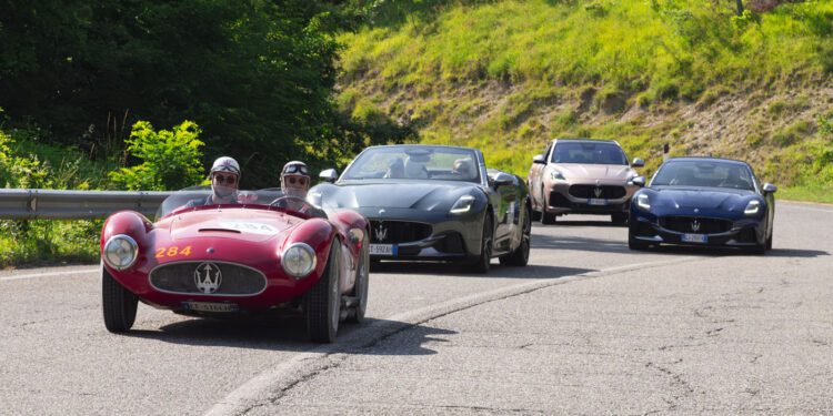 Four Maserati cars, including a vintage red race car reminiscent of the 1000 Miglia and three modern models, drive on a winding road bordered by greenery.